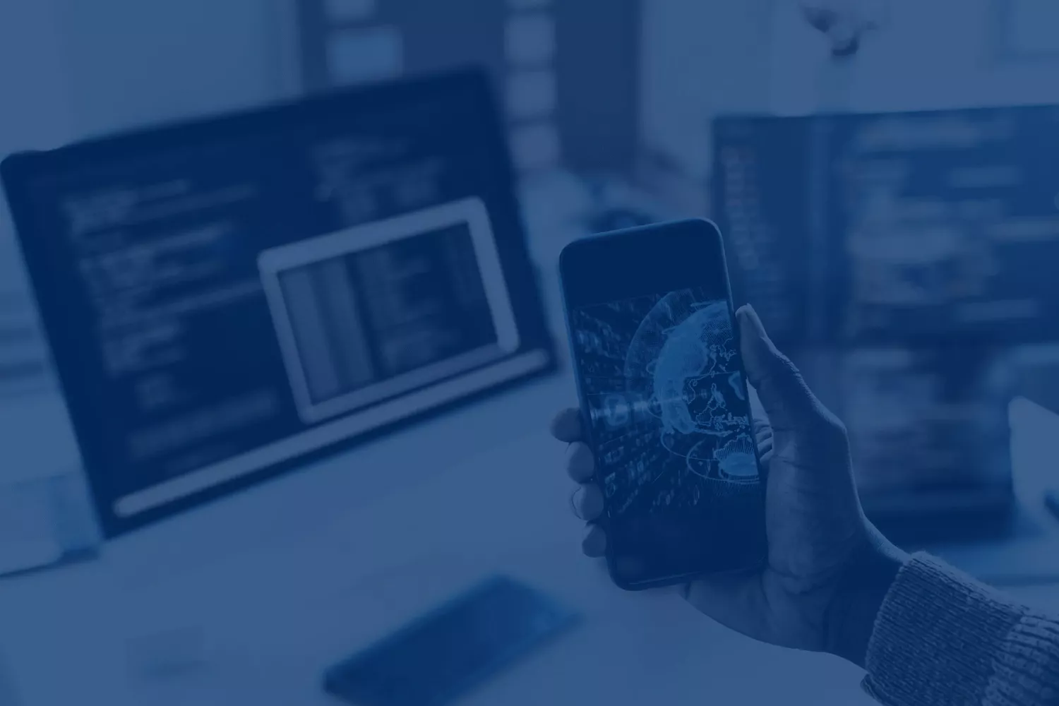 A hand holding a phone showing a data visualization, with code on the monitors behind it